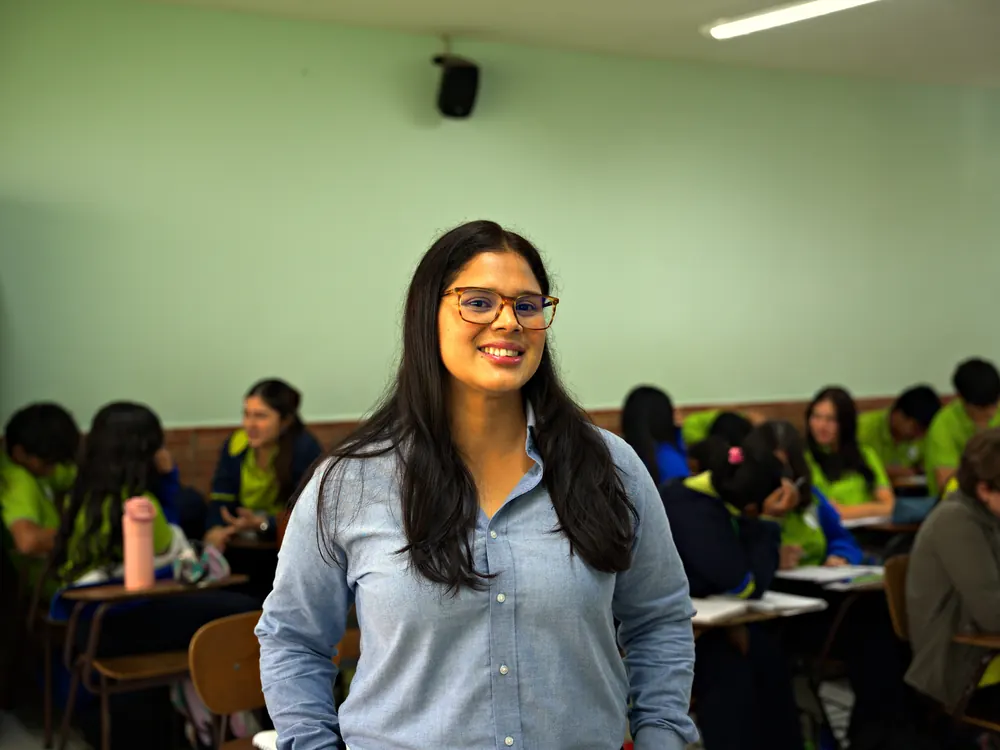 Docente de Secundaria sonriendo en su aula mientras los estudiantes trabajan en grupo